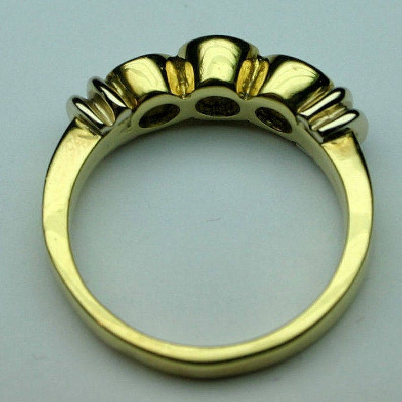 May include: A gold ring featuring a polished band and a setting that holds three circular stones. The metal has a smooth, reflective finish, and the design includes visible prongs securing each of the three stones along the top of the band.