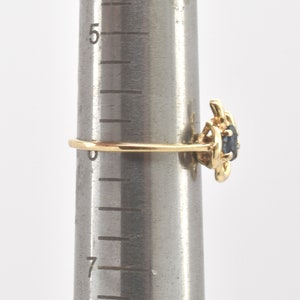 May include: A gold ring featuring a dark blue oval gemstone set in a decorative metal mounting. The ring is positioned on a metal ring mandrel marked with numerical sizing increments, showing the band resting near the zero mark on the scale.