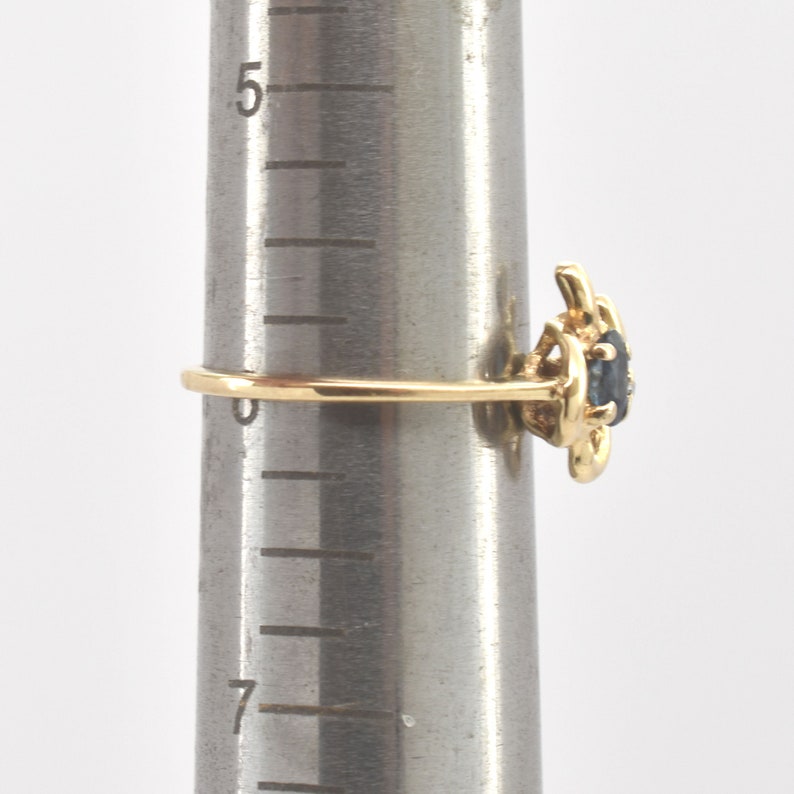 May include: A gold ring featuring a dark blue oval gemstone set in a decorative metal mounting. The ring is positioned on a metal ring mandrel marked with numerical sizing increments, showing the band resting near the zero mark on the scale.