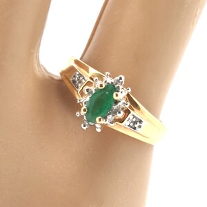May include: A gold ring featuring a central marquise-cut green gemstone surrounded by a cluster of small clear stones. The band is polished gold and includes additional small clear stones set into the shoulders on either side of the center.