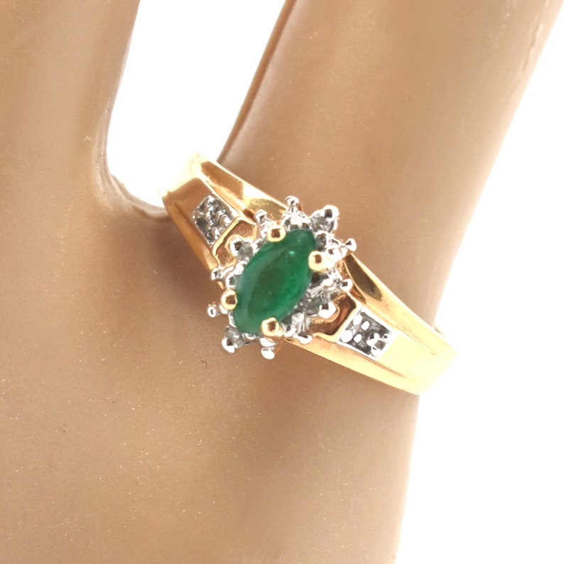May include: A gold ring featuring a central marquise-cut green gemstone surrounded by a cluster of small clear stones. The band is polished gold and includes additional small clear stones set into the shoulders on either side of the center.