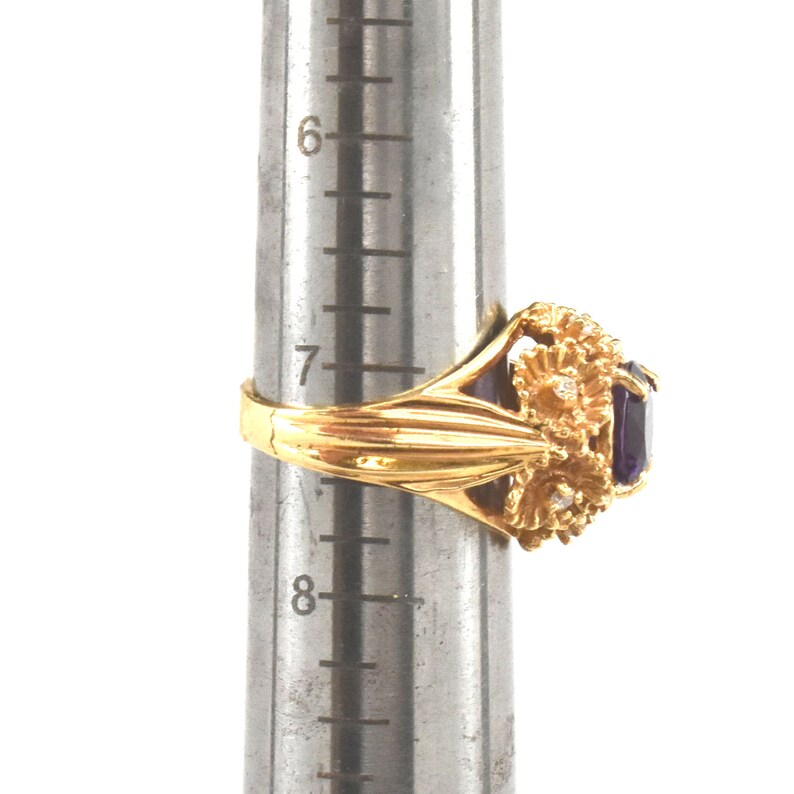 May include: A gold ring featuring a rectangular purple gemstone and floral detailing. The ring is on a ring sizer. The ring has small, round, clear stones set in the floral design.