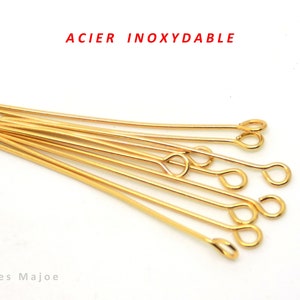 May include: Gold-colored eye pins with a loop at one end, scattered on a white surface. The text "ACIER INOXYDABLE" is in red at the top. The bottom left corner has the text "Perles Majoe".