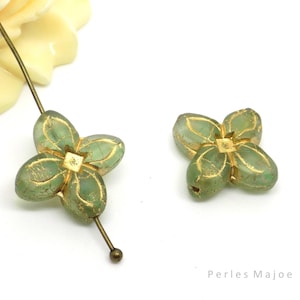 Könnte beinhalten: Zwei grüne Glasperlen in Blütenform mit goldenen Akzenten. Die Perlen haben ein vierblättriges Design und eine quadratische Mitte.