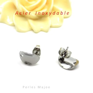 Peut inclure: Une paire de boucles d'oreilles à clous en acier inoxydable argenté avec une forme asymétrique lisse et arrondie. Chaque boucle comporte un petit trou circulaire près du bord pour la fixation. Le texte Acier Inoxydable est visible en rouge.