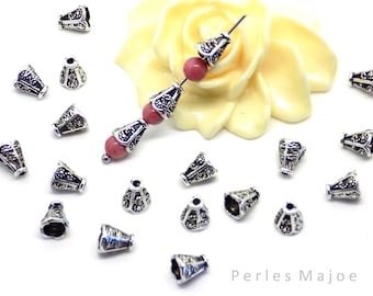 May include: A collection of silver-toned metal cone-shaped beads with intricate designs. The beads are scattered on a white surface with a pink flower in the background. The text "Perles Majoe" is visible in the bottom right corner of the image.