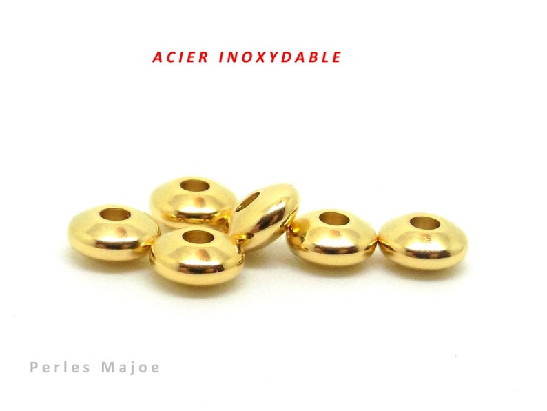 May include: Six gold-colored metal beads, each with a hole in the center. The beads are arranged in a row, with the text "ACIER INOXYDABLE" above them and "Perles Majoe" below them.