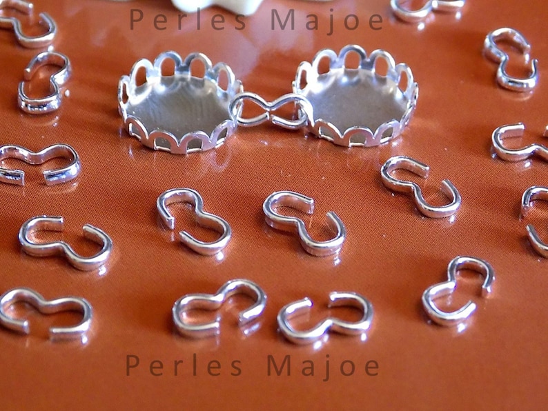 Puede incluir: Hallazgos de joyer&iacute;a de metal plateado, incluyendo un engaste festoneado y varios cierres en forma de S. Los hallazgos est&aacute;n dispuestos sobre una superficie marr&oacute;n. El texto "Perles Majoe" aparece dos veces en la imagen.