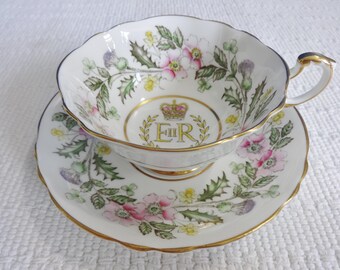 Könnte beinhalten: Eine weiße Teetasse und Untertasse mit goldfarbenem Rand. Tasse und Untertasse sind mit einem floralen Muster verziert, das rosa, gelbe und grüne Blumen zeigt. In der Mitte der Tasse befindet sich eine goldene Krone und die Buchstaben "E II R".