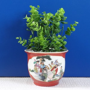 Op de afbeelding: Een rode en witte keramische pot met een geschilderd tafereel van mensen die een spel spelen. De pot is gevuld met groene, bladerige planten.