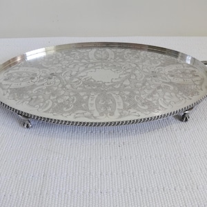 Könnte beinhalten: Ovale Serviertablett aus Silber mit aufwendigem Blumen- und Rankenmuster. Das Tablett hat zwei gebogene Griffe und steht auf kleinen Füßen. Der Rand hat ein gedrehtes Seildesign. Ein dekoratives Stück.