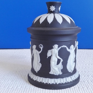 Pode incluir: Jarro de cerâmica preto com tampa branca e figuras brancas de mulheres dançando ao redor do jarro.