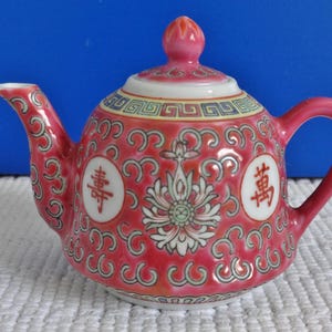 Może przedstawiać: Mały różowy porcelanowy czajniczek z kwiatowym wzorem i chińskimi znakami, które oznaczają "Szczęście" i "Dobrobyt".
