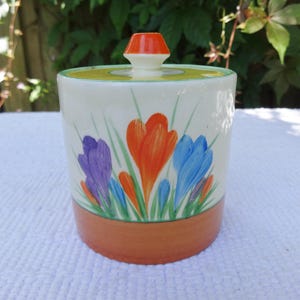 May include: A cylindrical ceramic jar with a lid featuring a hand-painted floral design of purple, orange, and blue crocus flowers with green stems. The base is a solid terracotta colour, and the lid has a small orange knob handle on top.