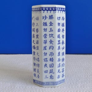 Op de afbeelding: Een witte porseleinen vaas met een zeshoekige vorm en blauwe en witte Chinese karakters. De vaas is versierd met een blauw en wit bloemenpatroon rond de boven- en onderkant.
