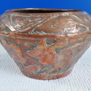 May include: A copper-coloured, handcrafted bowl with an embossed floral design. The bowl has a wide, rounded shape with a slightly flared base. The surface shows signs of age and wear, with areas of green oxidation. The bowl sits on a white surface.