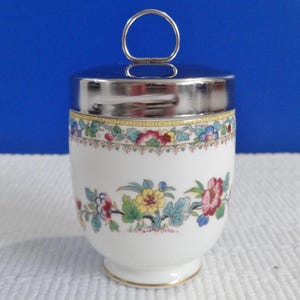 Könnte beinhalten: Eine weiße Porzellan-Eierbecher mit einem Blumenmuster und einem silbernen Deckel mit einem Griff. Der Eierbecher hat einen goldenen Rand um die Ober- und Unterseite.