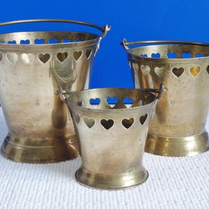 May include: Three brass buckets with heart-shaped cutouts. The buckets are nested inside each other, with the largest bucket at the bottom and the smallest bucket at the top.