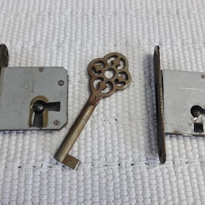 May include: Two silver door locks with a keyhole and a single ornate key with a flower-shaped head. The key is made of brass and has a worn finish.