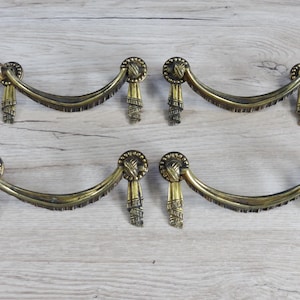 Könnte beinhalten: Satz von sechs antiken Messing-Schubladengriffen mit einem dekorativen Quasten-Design. Jeder Griff hat eine geschwungene Form mit einem Knotendetail oben und einer Fransenquaste, die darunter hängt.