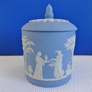 Op de afbeelding: Een blauwe en witte keramische pot met een deksel. De pot heeft een reliëfdesign met twee figuren in een tuinsetting.