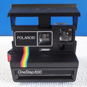 Peut inclure: Appareil photo instantané Polaroid OneStep 600 noir avec le nom de la marque en blanc. Une bande arc-en-ciel colorée longe le côté. L'appareil photo dispose d'un flash et d'un viseur. L'appareil photo est sur une surface blanche avec un fond bleu.