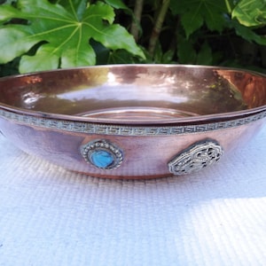 May include: A round, shallow copper bowl featuring a polished metallic finish. The exterior rim is decorated with a silver-toned Greek key pattern. Two distinct silver-toned ornaments are attached to the side, one featuring a blue stone.