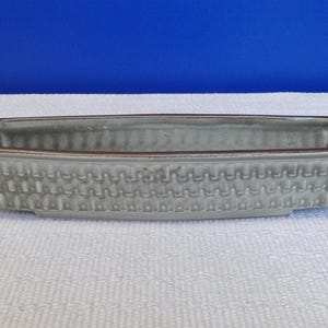 Może przedstawiać: Prostokątna szara ceramiczna donica z teksturowanym, przypominającym splot wzorem po bokach. Obręcz ma ciemnobrązowy kolor. Donica jest pusta i stoi na białej powierzchni z niebieskim tłem.