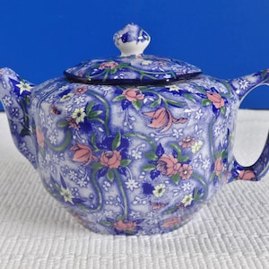 Puede incluir: Una tetera azul y blanca con un patrón floral. La tetera tiene una tapa y un asa. El patrón floral es en tonos de rosa, amarillo y verde.