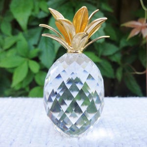Könnte beinhalten: Eine klare, facettierte Kristall-Ananas-Figur mit einem polierten goldfarbenen Metallaufsatz. Der Körper der Ananas weist ein rautenförmiges Schliffmuster auf, das Licht reflektiert, während die Metallblätter an der Krone glatt sind.