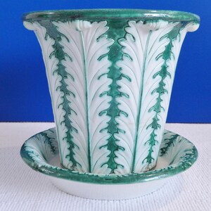 May include: A white ceramic planter with a green leafy design. The planter has a matching saucer.