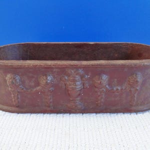 May include: An oval terracotta planter with a decorative design of figures and a central motif. The planter is a reddish-brown colour.