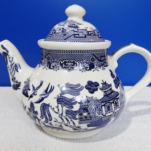 Puede incluir: Una tetera de cerámica blanca y azul con tapa. La tetera presenta un patrón Willow clásico con ilustraciones detalladas de pagodas, pájaros y paisajes. El asa y el pico son blancos.
