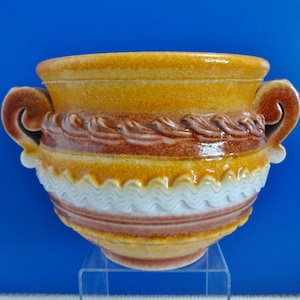 Può includere: Un piccolo vaso in ceramica marrone e bianco con due manici. Il vaso ha un design ondulato intorno al centro e un design a corda intorno alla parte superiore.