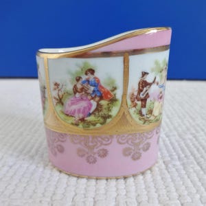 May include: A small, pink and gold porcelain creamer with a scalloped rim. The creamer is decorated with a floral pattern and scenes of people in 18th-century clothing.