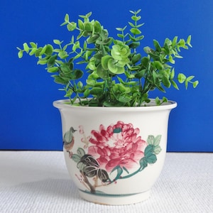 Puede incluir: Una maceta de cerámica blanca con un diseño floral que presenta una flor rosa y hojas verdes. Un pequeño pájaro está pintado en el lateral de la maceta. La maceta está llena de una planta verde de hojas.