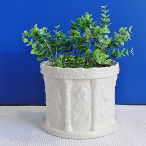 May include: A white ceramic plant pot with a detailed relief design, filled with vibrant green artificial foliage. The pot has a cylindrical shape with a decorative base and rim, set against a blue background.