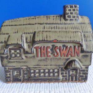 Può includere: Un edificio in ceramica in stile cottage con tetto di paglia e le parole "The Swan" in lettere marroni. L'edificio ha un camino e finestre.