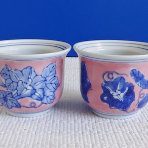 May include: Two small, pink and blue ceramic cups with a floral design. The cups have a white rim and a blue floral pattern on a pink background.