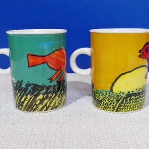 May include: Two ceramic mugs with white handles. One mug is green with a red bird design, the other is yellow with a red bird design. Both mugs have a white rim and a black and yellow grass design at the bottom.