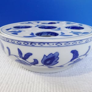 May include: A white ceramic flower frog bowl featuring a removable lid with multiple circular openings. The exterior and lid are decorated with a blue floral pattern and a geometric diamond border around the upper rim of the bowl.