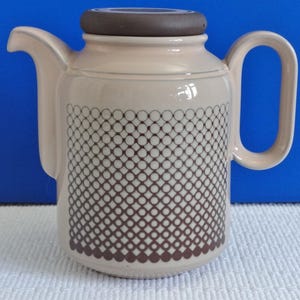 Peut inclure: Une théière en céramique beige avec un couvercle brun et un motif géométrique de cercles bruns sur le corps. La théière a une poignée et un bec.