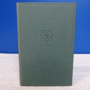May include: A teal-covered hardcover book. The cover has a decorative emblem above the name "RUSKIN". The book is upright against a blue backdrop. The book's spine is visible.