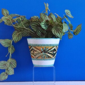 Könnte beinhalten: Ein weißer Keramik-Blumentopf mit einem geometrischen Muster in Gold, Braun und Türkis, der eine grüne Pflanze mit gestreiften Blättern enthält. Der Topf steht auf einem transparenten Ständer vor blauem Hintergrund.