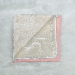 May include: A baby blanket with a soft, plush cream-colored interior and a light pink exterior. The blanket is trimmed with a white pom-pom border. The blanket is folded, revealing the cream-colored interior.