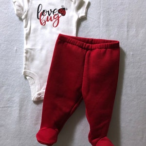 May include: A white baby onesie with the text "love bug" and a red ladybug graphic. The onesie is paired with red footed pants.