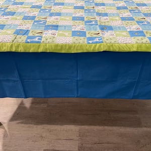 May include: A blue tablecloth with a green and white patterned table cover on top. The table cover has a satin trim.