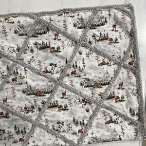 May include: A quilted blanket featuring a woodland scene with deer, trees, and birds in shades of brown, green, and gray. The blanket has a gray, textured border and diamond-shaped stitching.