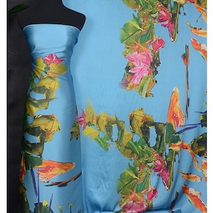 May include: Blue satin fabric with a colorful abstract floral print. The fabric is draped over a mannequin and a table. The fabric has a bright blue background with yellow, orange, green, pink, and white flowers.