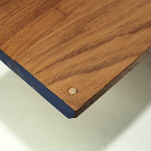 May include: Close-up of a wooden table corner. The tabletop is a warm brown with visible wood grain. The edge is painted a deep blue, and a small, round, brass-colored fastener is visible.